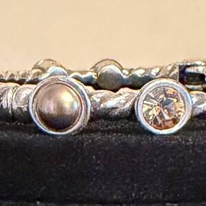 May include: A set of silver-toned bangles with a twisted rope design. Each bangle features decorative accents, including round, bezel-set stones in shades of brown and gold. The bangles are displayed on a black surface.