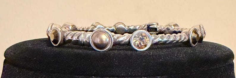 May include: A set of silver-toned bangles with a twisted rope design. Each bangle features decorative accents, including round, bezel-set stones in shades of brown and gold. The bangles are displayed on a black surface.