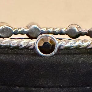 May include: Two silver-toned bangles with a twisted rope design. Each bangle features alternating round and faceted, light brown gemstones. The bangles are displayed on a black surface against a neutral background.