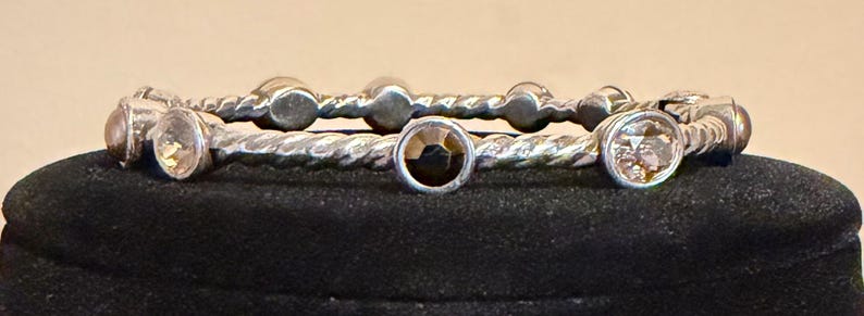 May include: Two silver-toned bangles with a twisted rope design. Each bangle features alternating round and faceted, light brown gemstones. The bangles are displayed on a black surface against a neutral background.
