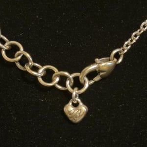 May include: Silver-toned necklace with a heart-shaped charm. The charm has an engraved design and hangs from a small chain. The necklace features a lobster clasp and a series of interconnected circular links.