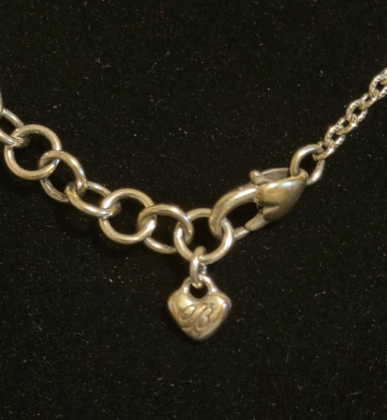 May include: Silver-toned necklace with a heart-shaped charm. The charm has an engraved design and hangs from a small chain. The necklace features a lobster clasp and a series of interconnected circular links.