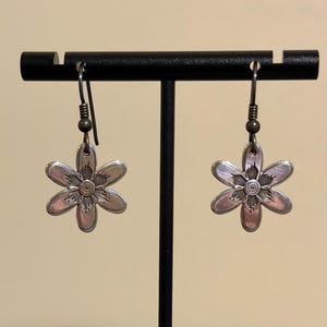 Może przedstawiać: Para srebrnych kolczyk&oacute;w w kształcie kwiat&oacute;w. Każdy kolczyk ma sześciopłatkowy wz&oacute;r kwiatu ze spiralnym środkiem. Kolczyki wiszą na prostym haczyku i są prezentowane na czarnym stojaku.