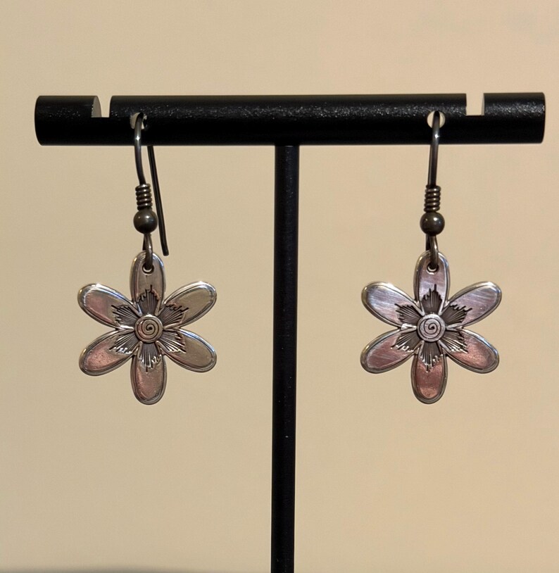 Może przedstawiać: Para srebrnych kolczyk&oacute;w w kształcie kwiat&oacute;w. Każdy kolczyk ma sześciopłatkowy wz&oacute;r kwiatu ze spiralnym środkiem. Kolczyki wiszą na prostym haczyku i są prezentowane na czarnym stojaku.