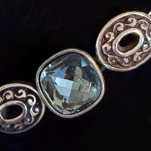 May include: Close-up of a silver bracelet featuring three decorative elements. A square, faceted, pale blue gemstone is the central piece. The other two pieces are oval-shaped with an open centre and ornate silver scrollwork. The background is black.
