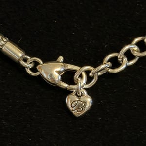 Puede incluir: Primer plano de un cierre de collar plateado con un dije en forma de coraz&oacute;n. El dije tiene la letra "B" grabada. El collar tiene un dise&ntilde;o de eslabones de cadena y una secci&oacute;n trenzada.