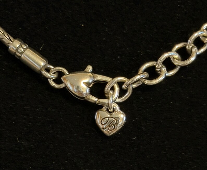 Puede incluir: Primer plano de un cierre de collar plateado con un dije en forma de coraz&oacute;n. El dije tiene la letra "B" grabada. El collar tiene un dise&ntilde;o de eslabones de cadena y una secci&oacute;n trenzada.