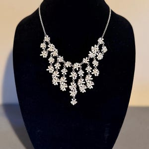 May include: A silver necklace and matching earrings set. The necklace has a cascade of small, white flower pendants. The earrings are small flower studs. The jewellery is displayed on a black velvet bust.