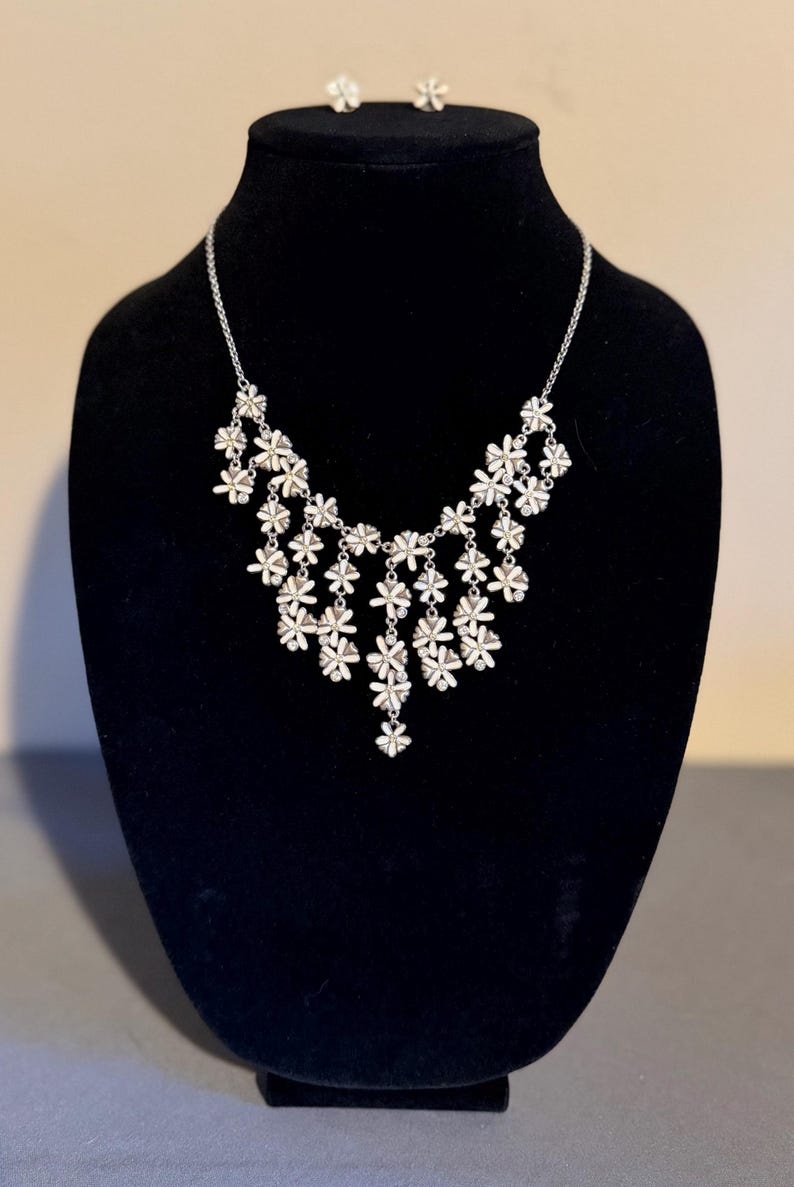 May include: A silver necklace and matching earrings set. The necklace has a cascade of small, white flower pendants. The earrings are small flower studs. The jewellery is displayed on a black velvet bust.