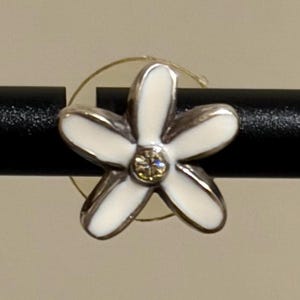 May include: A silver flower-shaped earring with white enamel petals and a central yellow gemstone. The earring is set against a black rod and a neutral background.