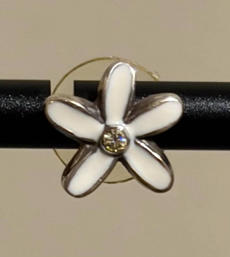 May include: A silver flower-shaped earring with white enamel petals and a central yellow gemstone. The earring is set against a black rod and a neutral background.