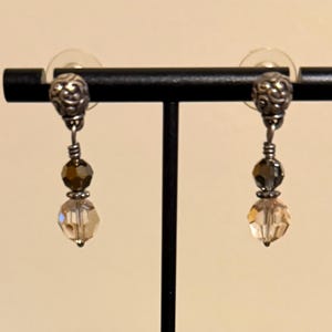 May include: A pair of dangle earrings displayed on a black stand. Each earring features a silver-toned top with a decorative design, a dark brown bead, a gray bead, and a faceted, light-colored crystal at the bottom.