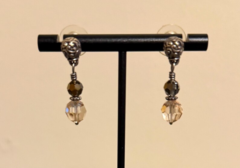 May include: A pair of dangle earrings displayed on a black stand. Each earring features a silver-toned top with a decorative design, a dark brown bead, a gray bead, and a faceted, light-colored crystal at the bottom.