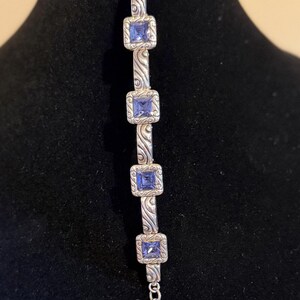 Puede incluir: Pulsera plateada con piedras preciosas azules cuadradas engastadas en marcos decorativos. La pulsera tiene un dise&ntilde;o ornamentado con una extensi&oacute;n de cadena y un dije en forma de coraz&oacute;n. La pulsera se exhibe en un maniqu&iacute; negro.