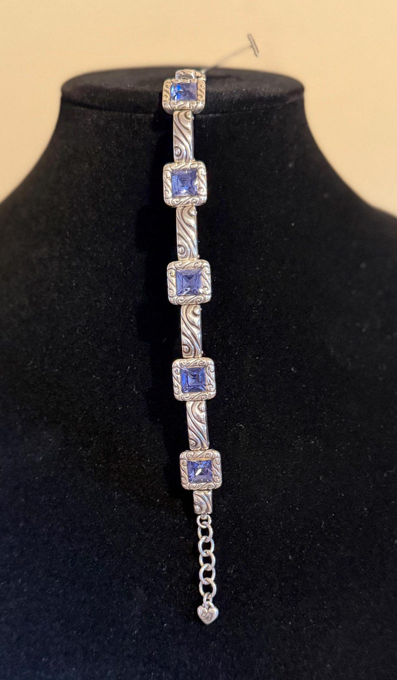Puede incluir: Pulsera plateada con piedras preciosas azules cuadradas engastadas en marcos decorativos. La pulsera tiene un dise&ntilde;o ornamentado con una extensi&oacute;n de cadena y un dije en forma de coraz&oacute;n. La pulsera se exhibe en un maniqu&iacute; negro.