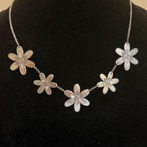 Może przedstawiać: Srebrny naszyjnik z pięcioma zawieszkami w kształcie stokrotek. Każdy kwiat ma szczeg&oacute;łowe płatki i spiralny środek. Naszyjnik jest prezentowany na czarnym tle, podkreślając design biżuterii.