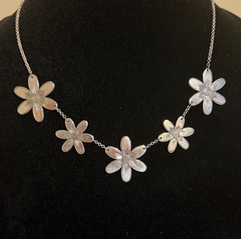 Może przedstawiać: Srebrny naszyjnik z pięcioma zawieszkami w kształcie stokrotek. Każdy kwiat ma szczeg&oacute;łowe płatki i spiralny środek. Naszyjnik jest prezentowany na czarnym tle, podkreślając design biżuterii.