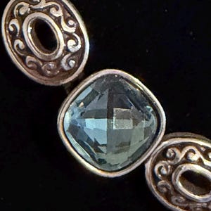 May include: Close-up of a silver bracelet featuring a square, faceted, light blue gemstone. The bracelet has two oval silver links with a decorative swirl design on either side of the gemstone.