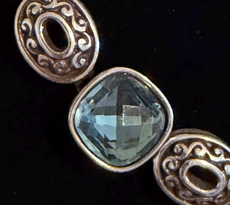 May include: Close-up of a silver bracelet featuring a square, faceted, light blue gemstone. The bracelet has two oval silver links with a decorative swirl design on either side of the gemstone.