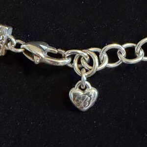 May include: Close-up of a silver bracelet featuring a heart-shaped charm. The charm has an engraved design and hangs from a chain link. The bracelet is set against a dark background.