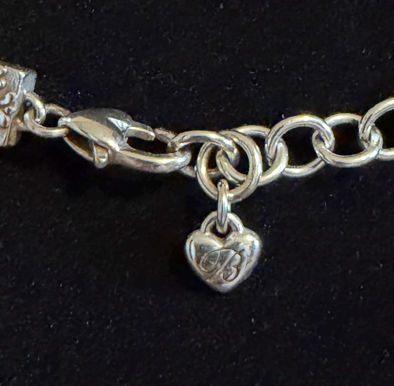 May include: Close-up of a silver bracelet featuring a heart-shaped charm. The charm has an engraved design and hangs from a chain link. The bracelet is set against a dark background.