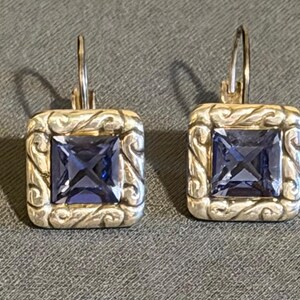 Puede incluir: Un par de pendientes colgantes cuadrados con un marco de metal plateado y un centro de piedra preciosa azul oscuro de corte cuadrado. El marco presenta un dise&ntilde;o ornamental y arremolinado. Cierre de palanca.