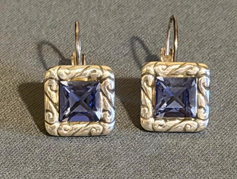 Puede incluir: Un par de pendientes colgantes cuadrados con un marco de metal plateado y un centro de piedra preciosa azul oscuro de corte cuadrado. El marco presenta un dise&ntilde;o ornamental y arremolinado. Cierre de palanca.