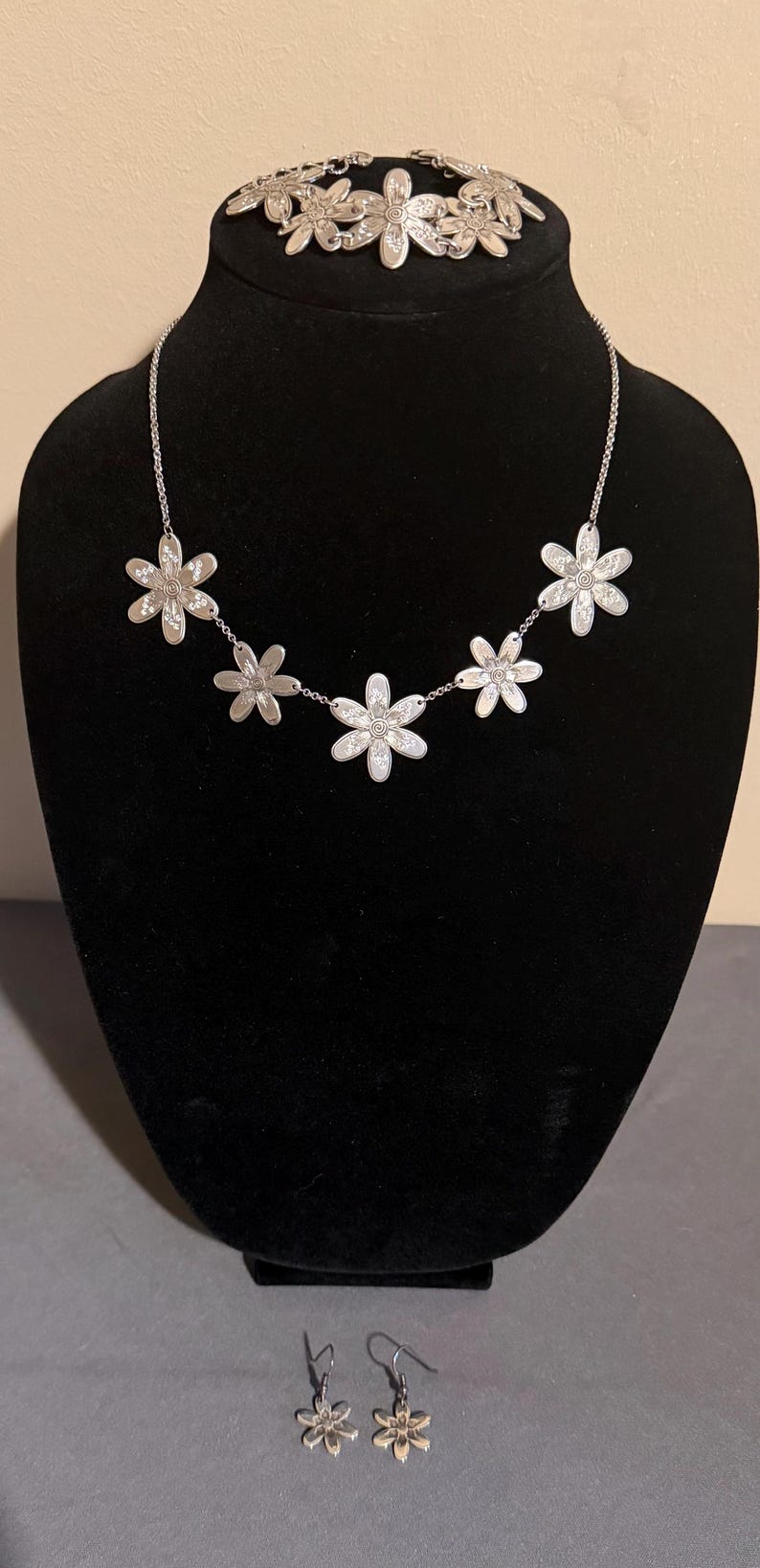 Może przedstawiać: Srebrny zestaw biżuterii w kwiaty, w tym naszyjnik, bransoletka i kolczyki, prezentowany na czarnym manekinie. Naszyjnik ma pięć kwiat&oacute;w przypominających stokrotki. Bransoletka znajduje się na g&oacute;rze manekina.