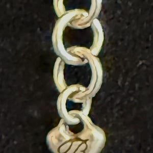 May include: Close-up of a silver chain with oval links. The chain ends with a heart-shaped charm, engraved with the cursive letter "B". The background is a dark, solid colour.