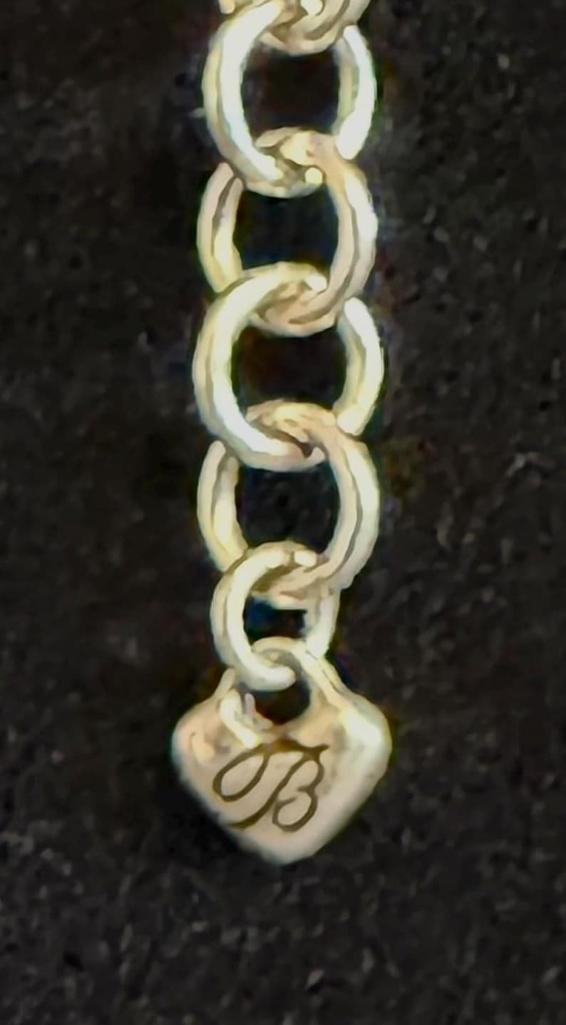 May include: Close-up of a silver chain with oval links. The chain ends with a heart-shaped charm, engraved with the cursive letter "B". The background is a dark, solid colour.