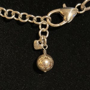 Puede incluir: Primer plano de un cierre y cadena de collar plateados. Los eslabones de la cadena son grandes y redondos. Un peque&ntilde;o dije con un dise&ntilde;o floral cuelga del cierre, junto con un dije de bola decorativa.