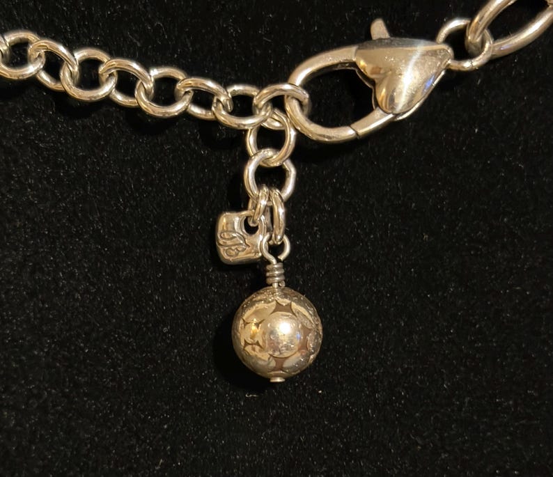 Puede incluir: Primer plano de un cierre y cadena de collar plateados. Los eslabones de la cadena son grandes y redondos. Un peque&ntilde;o dije con un dise&ntilde;o floral cuelga del cierre, junto con un dije de bola decorativa.