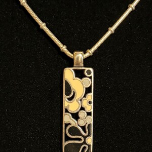 K&ouml;nnte beinhalten: Silberfarbene Halskette mit rechteckigem Anh&auml;nger mit floralem Design in Schwarz, Gelb und Grau. Der Anh&auml;nger h&auml;ngt an einer filigranen Kette mit kleinen, runden Perlen. Der Hintergrund ist schwarz.