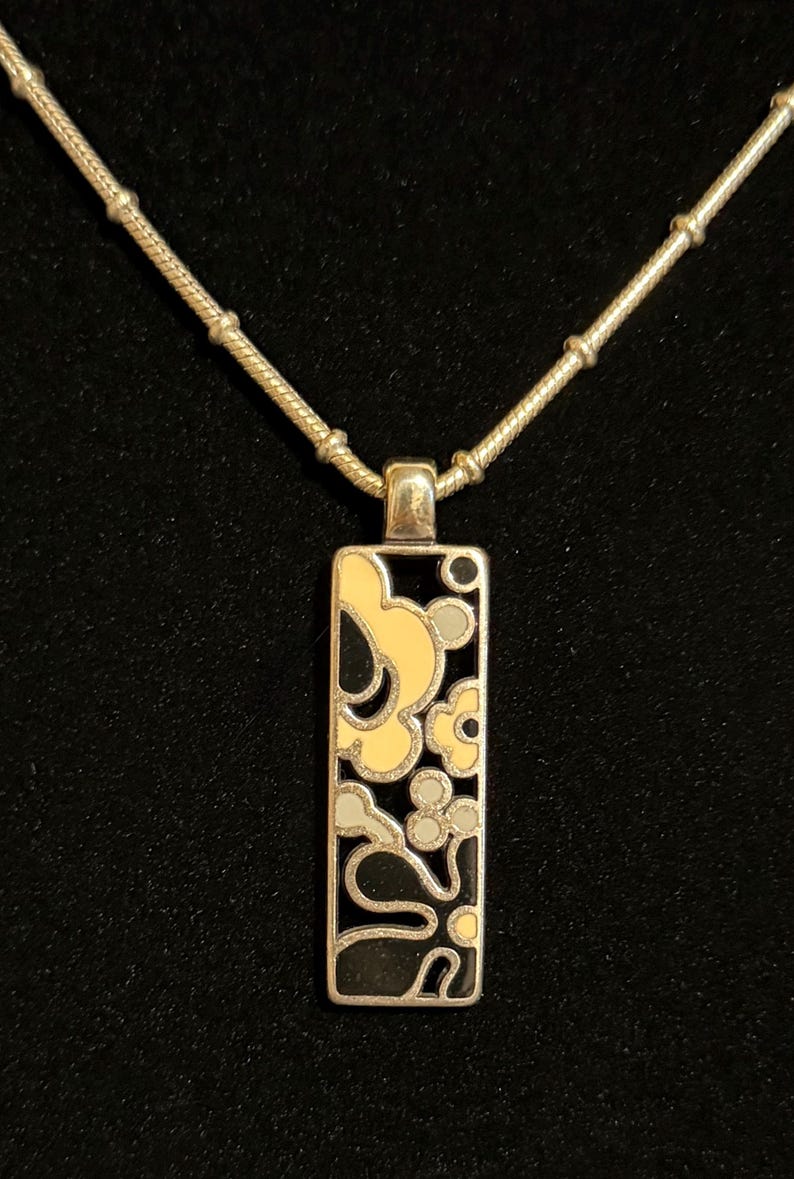 K&ouml;nnte beinhalten: Silberfarbene Halskette mit rechteckigem Anh&auml;nger mit floralem Design in Schwarz, Gelb und Grau. Der Anh&auml;nger h&auml;ngt an einer filigranen Kette mit kleinen, runden Perlen. Der Hintergrund ist schwarz.