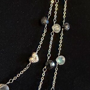 May include: A multi-strand necklace with silver chains and various beads. The necklace features clear, faceted beads, dark grey faceted beads, and silver heart-shaped and round beads. The background is a solid black.