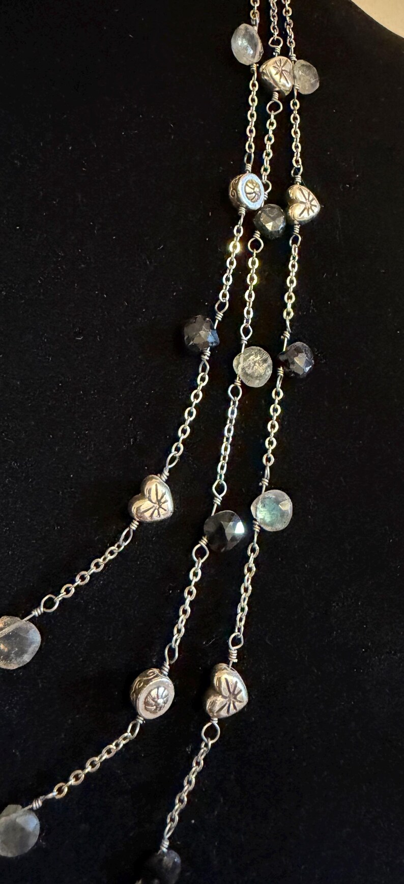 May include: A multi-strand necklace with silver chains and various beads. The necklace features clear, faceted beads, dark grey faceted beads, and silver heart-shaped and round beads. The background is a solid black.