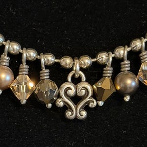 May include: Close-up of a silver necklace with a heart-shaped pendant and dangling beads. The necklace features a mix of round, faceted, and pearl-like beads in silver, brown, and amber colors. The background is black.
