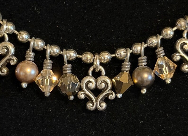 May include: Close-up of a silver necklace with a heart-shaped pendant and dangling beads. The necklace features a mix of round, faceted, and pearl-like beads in silver, brown, and amber colors. The background is black.