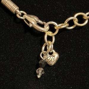 K&ouml;nnte beinhalten: Nahaufnahme eines silberfarbenen Halskettenverschlusses und einer Kette. Ein kleiner herzf&ouml;rmiger Anh&auml;nger mit dem Buchstaben "B" und zwei dunkelgrauen Kristallperlen baumeln an der Kette. Der Hintergrund ist schwarz.