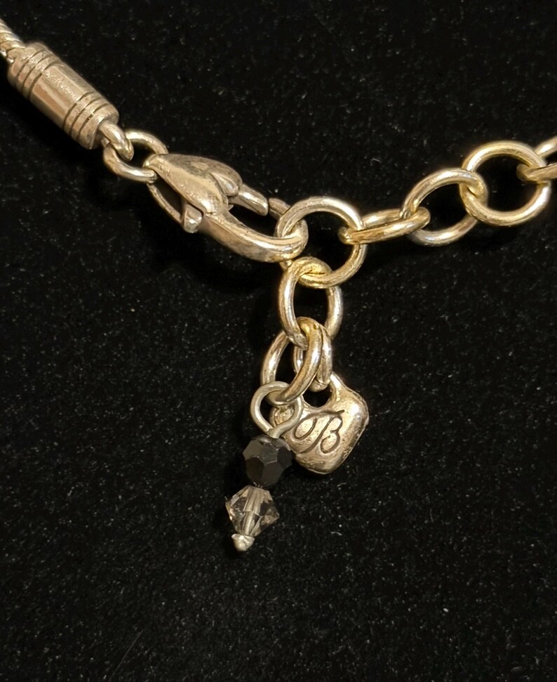 K&ouml;nnte beinhalten: Nahaufnahme eines silberfarbenen Halskettenverschlusses und einer Kette. Ein kleiner herzf&ouml;rmiger Anh&auml;nger mit dem Buchstaben "B" und zwei dunkelgrauen Kristallperlen baumeln an der Kette. Der Hintergrund ist schwarz.