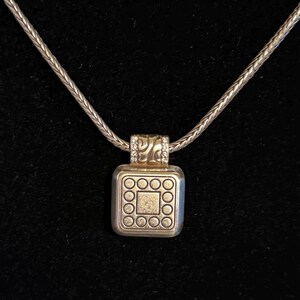 Puede incluir: Collar plateado con un colgante cuadrado. El colgante presenta un cuadrado central rodeado de c&iacute;rculos y una secci&oacute;n superior decorativa con peque&ntilde;as piedras transparentes. La cadena tiene un dise&ntilde;o tejido y el fondo es negro.