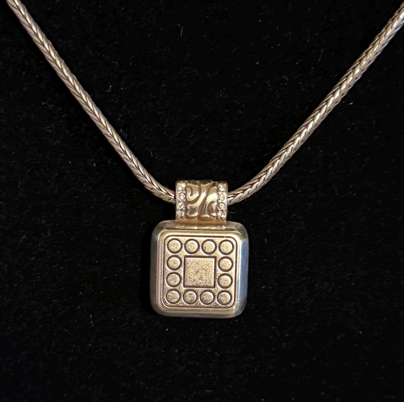 Puede incluir: Collar plateado con un colgante cuadrado. El colgante presenta un cuadrado central rodeado de c&iacute;rculos y una secci&oacute;n superior decorativa con peque&ntilde;as piedras transparentes. La cadena tiene un dise&ntilde;o tejido y el fondo es negro.