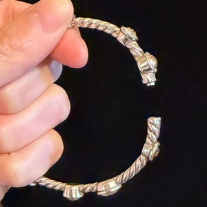 May include: Silver-toned bracelet with a twisted rope design. The bracelet features decorative embellishments and is held open, showcasing its intricate details. The background is a solid black.