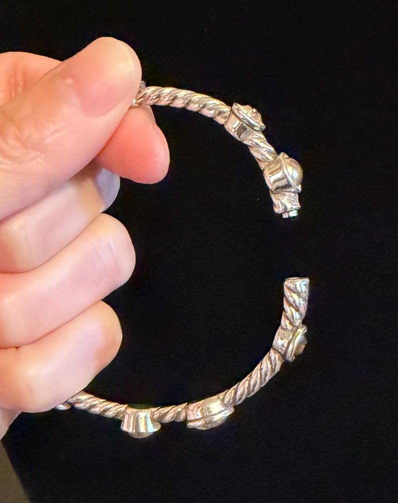 May include: Silver-toned bracelet with a twisted rope design. The bracelet features decorative embellishments and is held open, showcasing its intricate details. The background is a solid black.