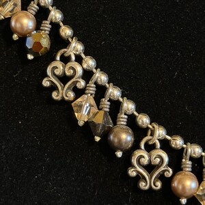 May include: Close-up of a silver-toned necklace with decorative heart-shaped pendants and various beads. The beads include faceted crystals and round, pearl-like beads in shades of brown and gold. The necklace is on a black background.