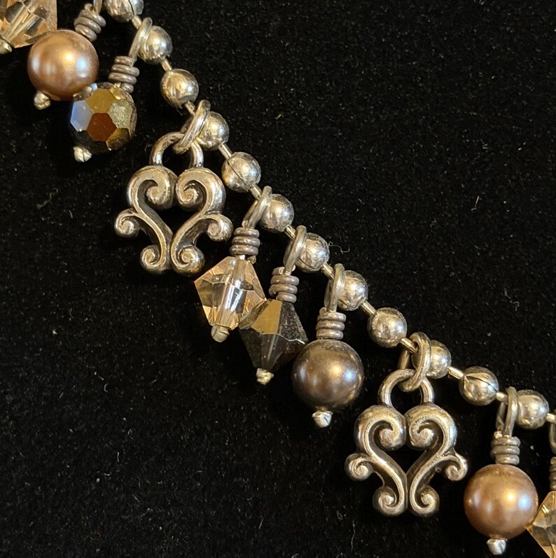 May include: Close-up of a silver-toned necklace with decorative heart-shaped pendants and various beads. The beads include faceted crystals and round, pearl-like beads in shades of brown and gold. The necklace is on a black background.