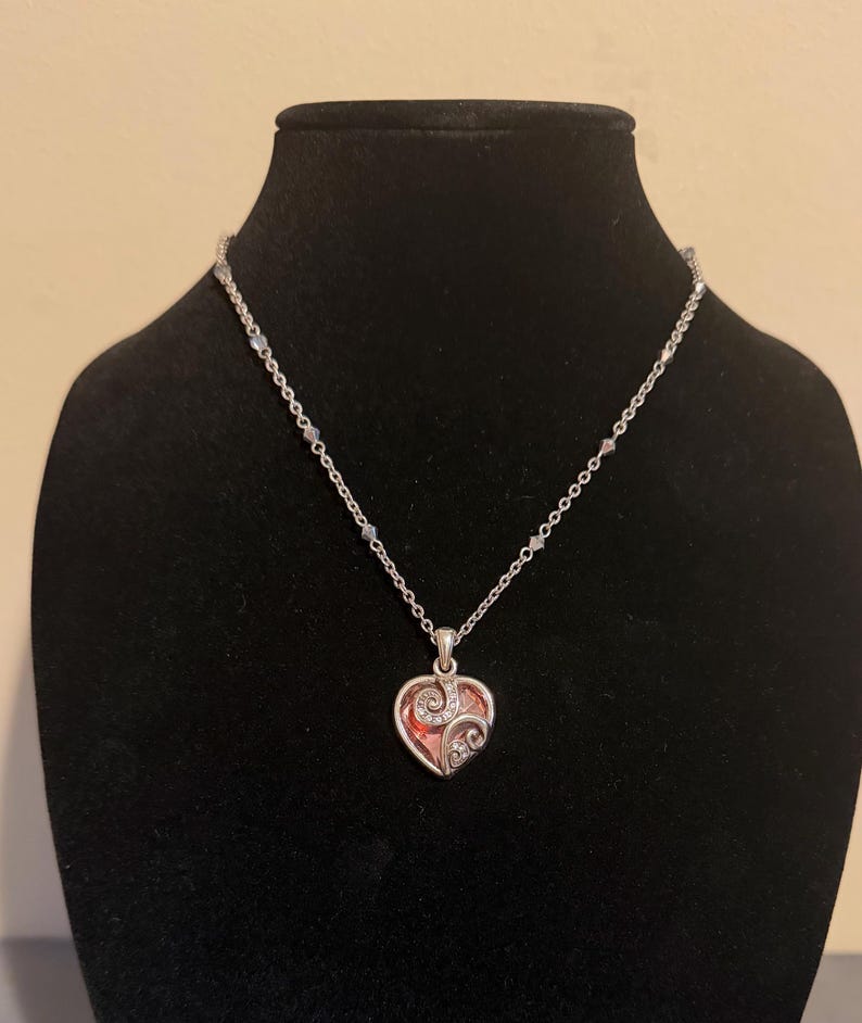 May include: Silver-toned necklace with a heart-shaped pendant. The pendant features a red center, clear crystals, and a decorative swirl design. The necklace is displayed on a black mannequin.