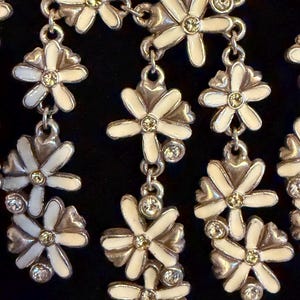 May include: A silver necklace with white enamel daisy flower pendants, each with a yellow centre and clear crystal details. The floral design is linked by a delicate chain, creating an elegant accessory. The necklace is suitable for various occasions.
