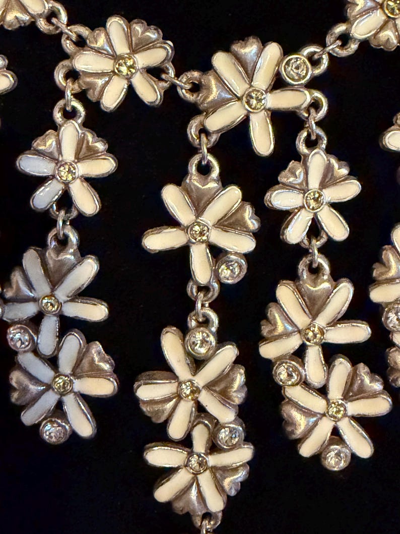 May include: A silver necklace with white enamel daisy flower pendants, each with a yellow centre and clear crystal details. The floral design is linked by a delicate chain, creating an elegant accessory. The necklace is suitable for various occasions.