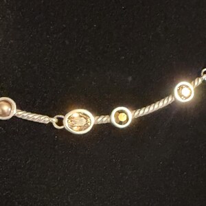 May include: Silver-toned necklace with a twisted rope chain. The necklace features a series of bezel-set, oval and round gemstones in shades of gold and amber. The gemstones are evenly spaced along the chain.